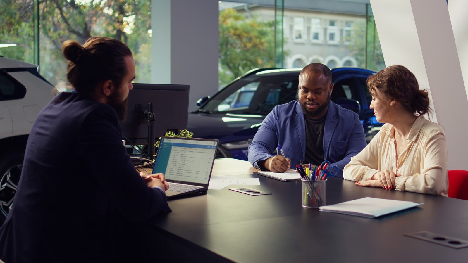 Car dealership salesman sitting with clients at table, giving them contract to sign